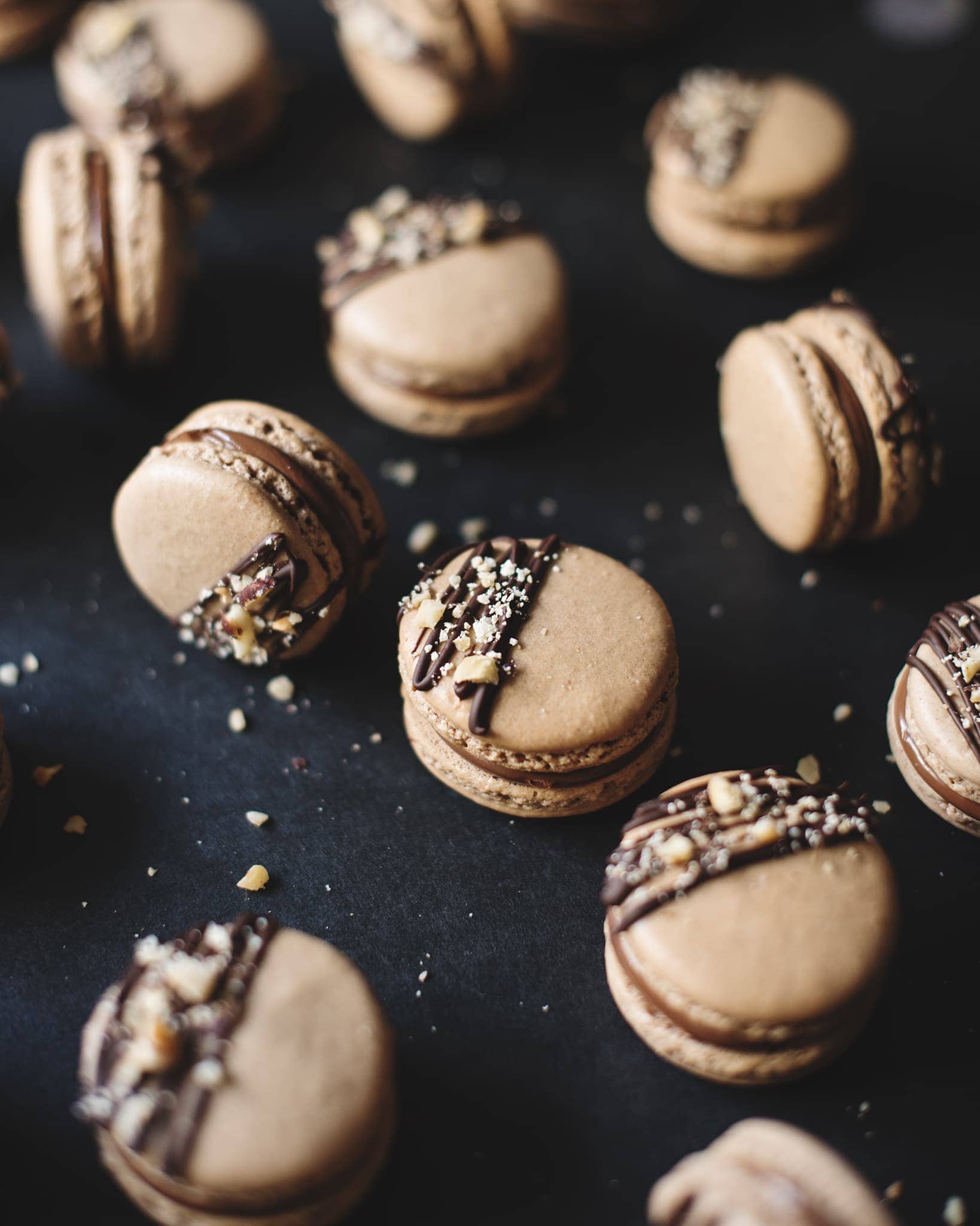 Ferrero Rocher macarons topped with dark chocolate drizzle and crushed hazelnuts scattered on a black background