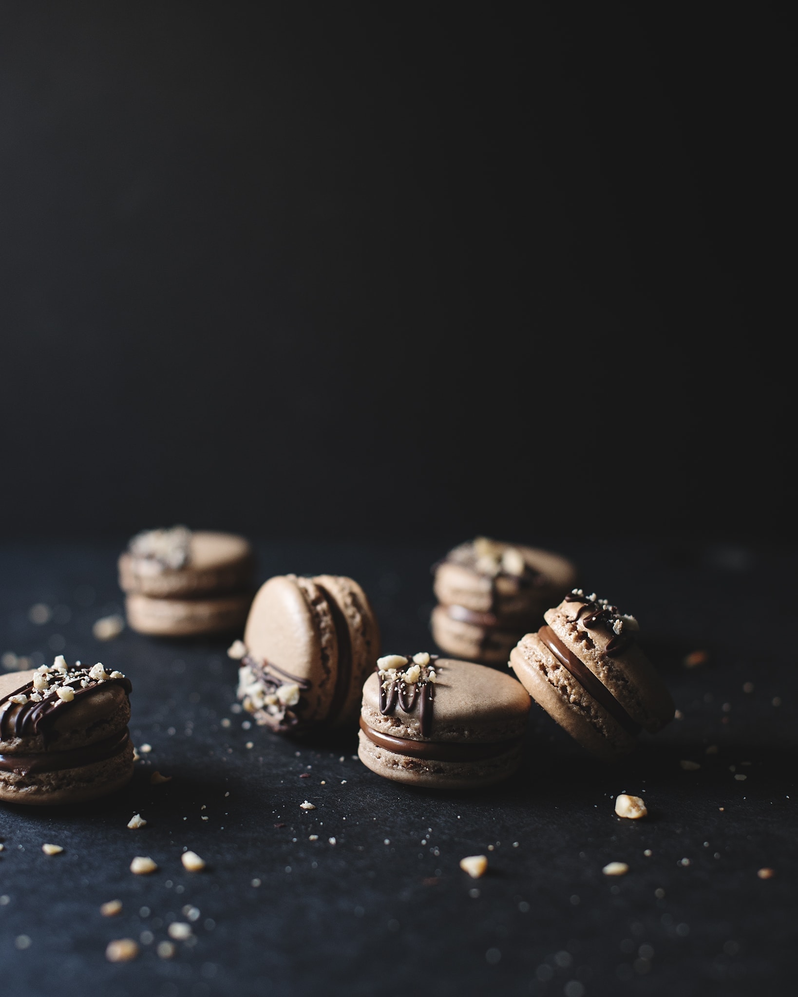 Ferrero Rocher macarons topped with dark chocolate drizzle and crushed hazelnuts scattered on a black background
