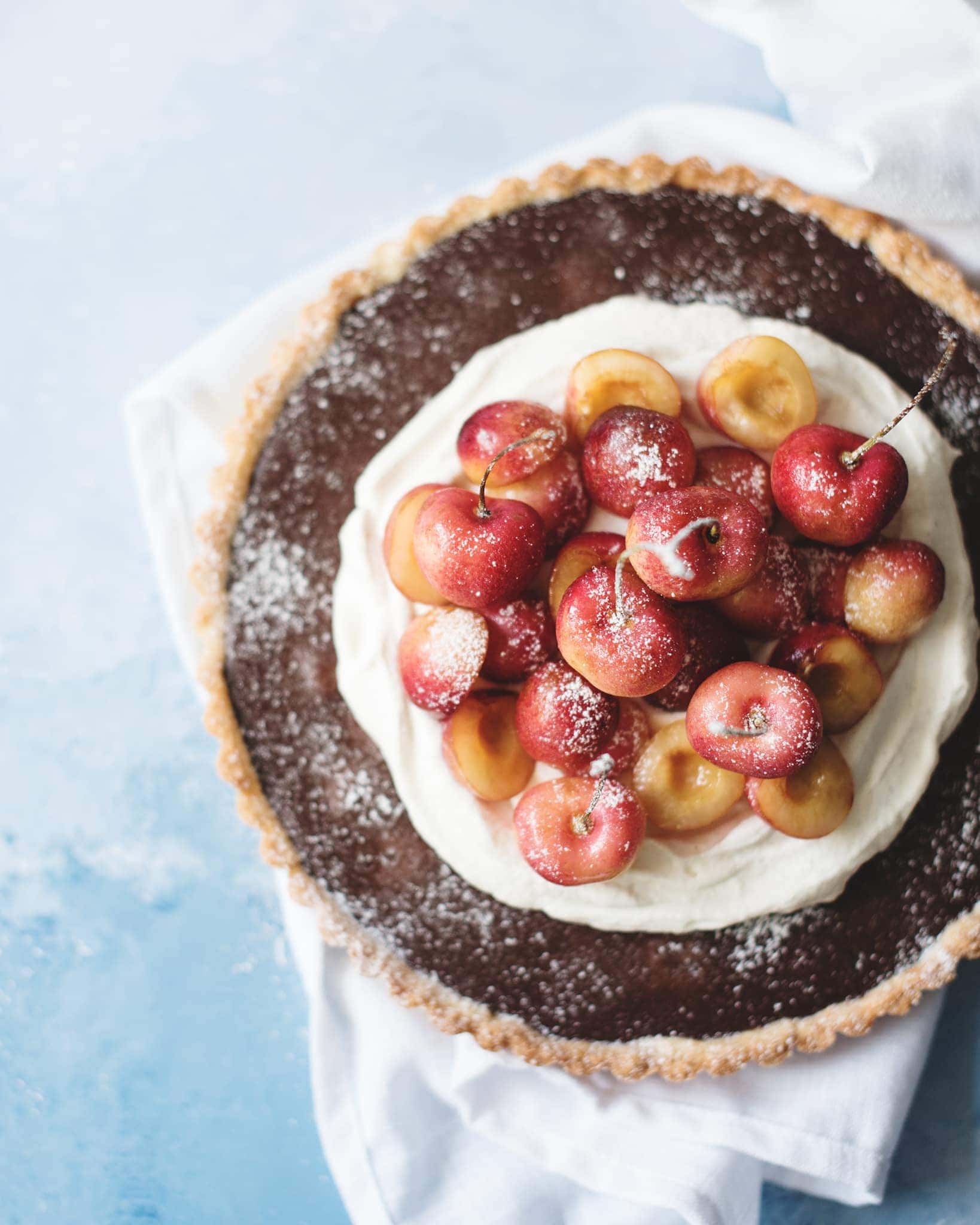 Overhead shot of chocolate cherry tart with cardamom cream on white napkin and blue background