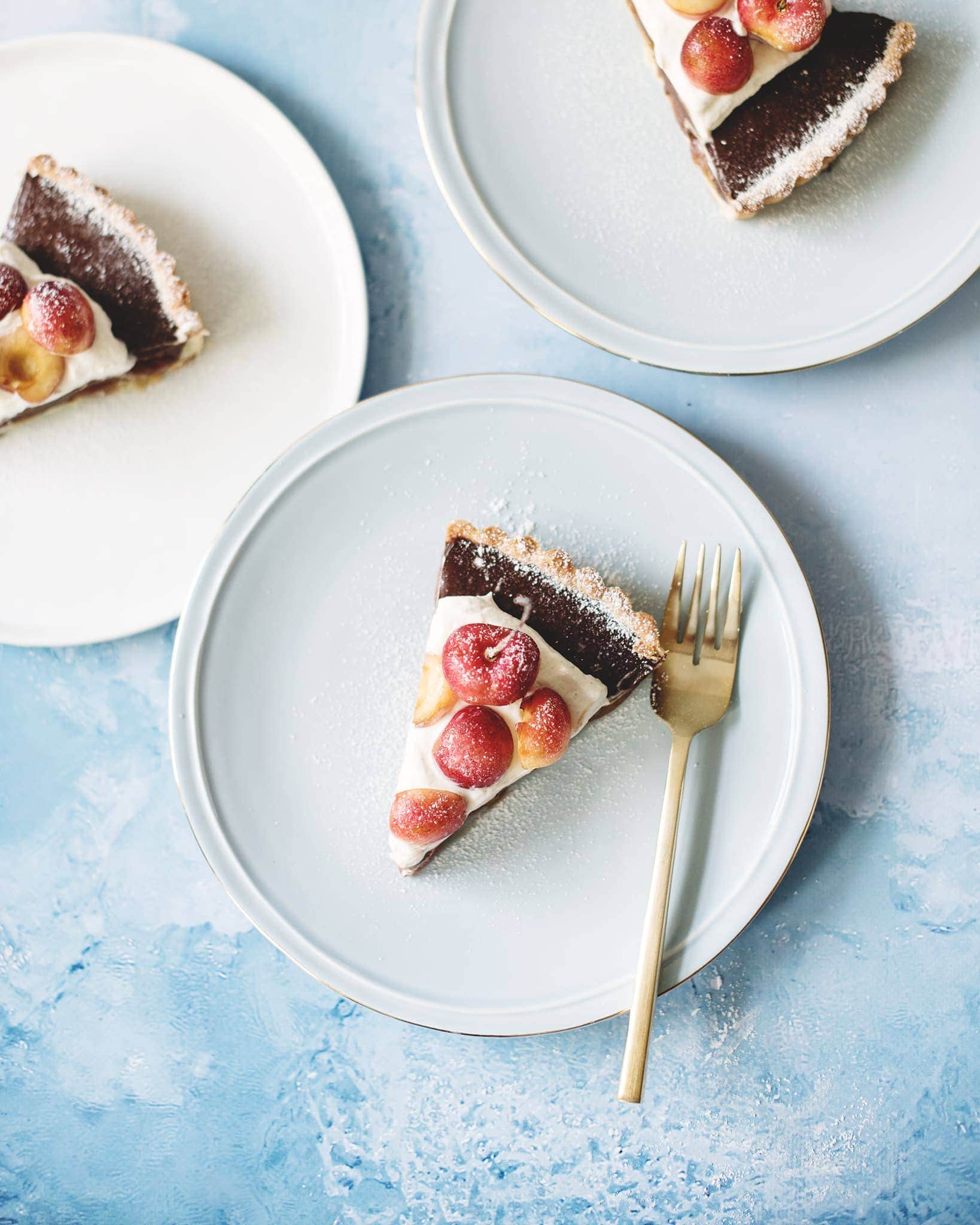 Overhead shot of slices of chocolate cherry tart with cardamom cream on blue plates and blue background