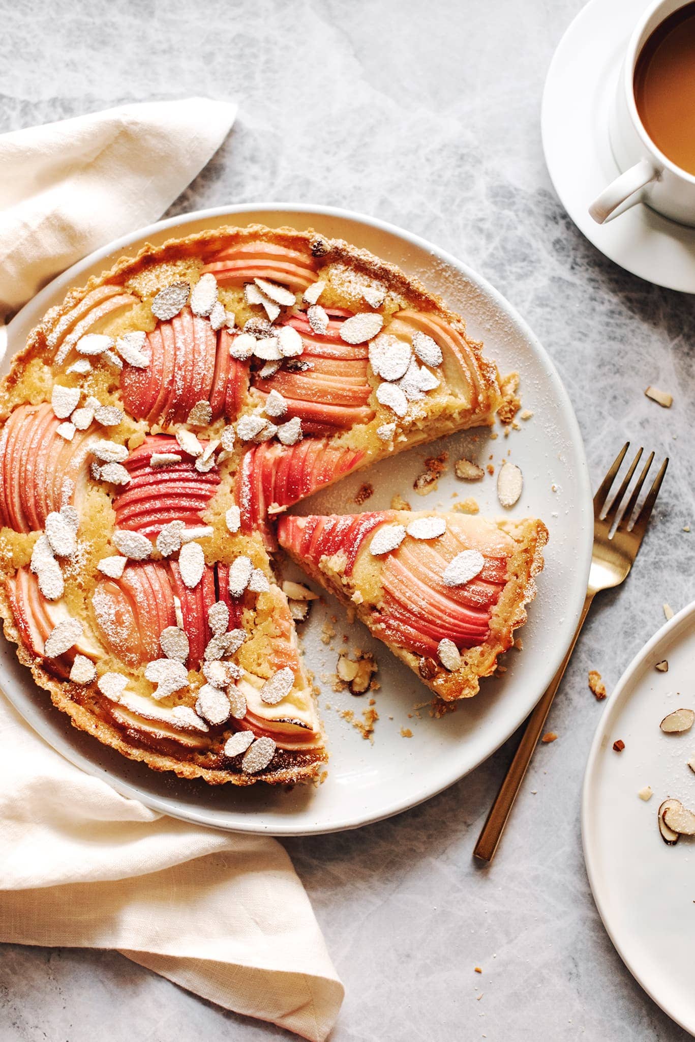 A slice cut out of an apple frangipane tart on a grey background with a cup of coffee