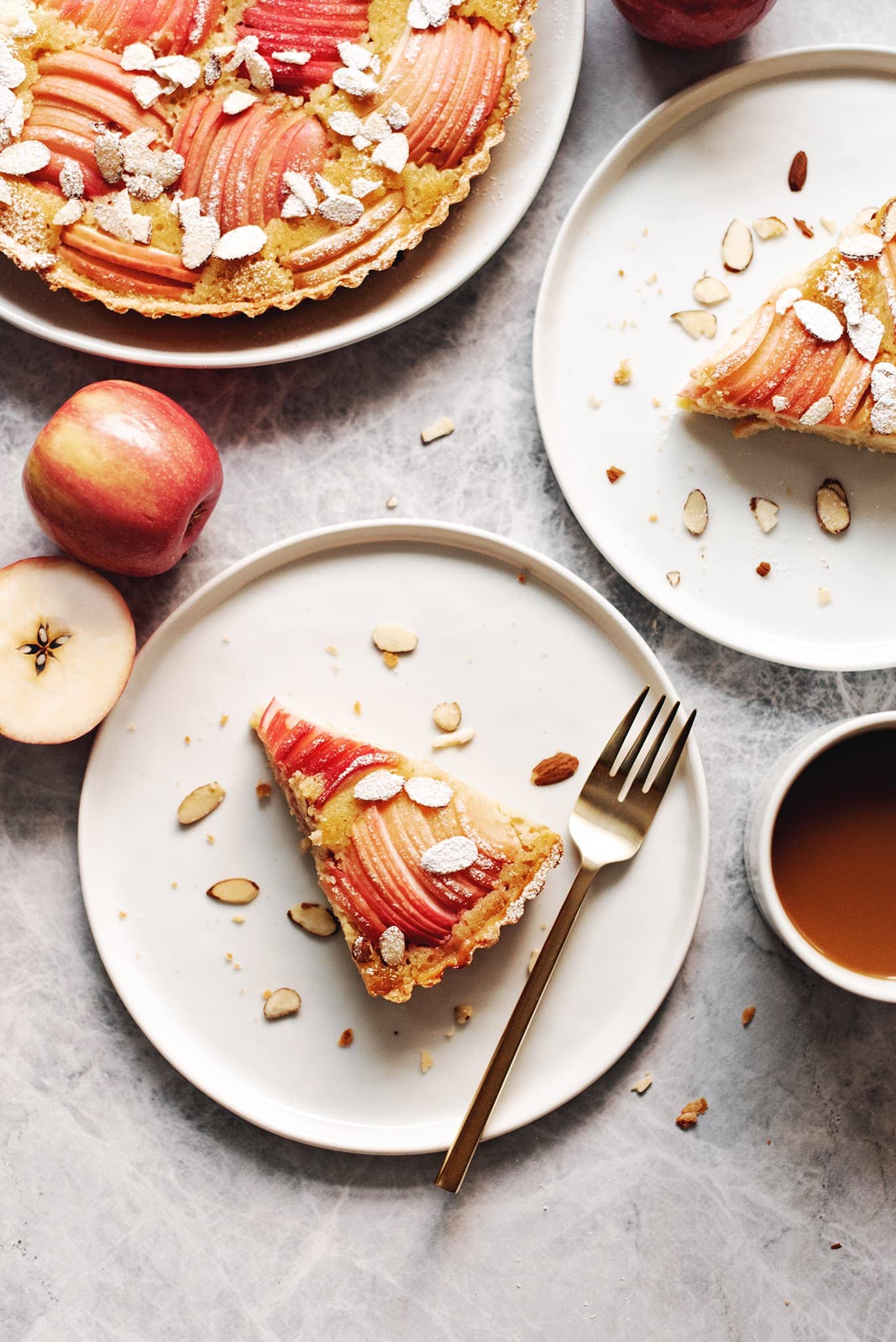 Two slices of apple frangipane tart on white plates with gold forks