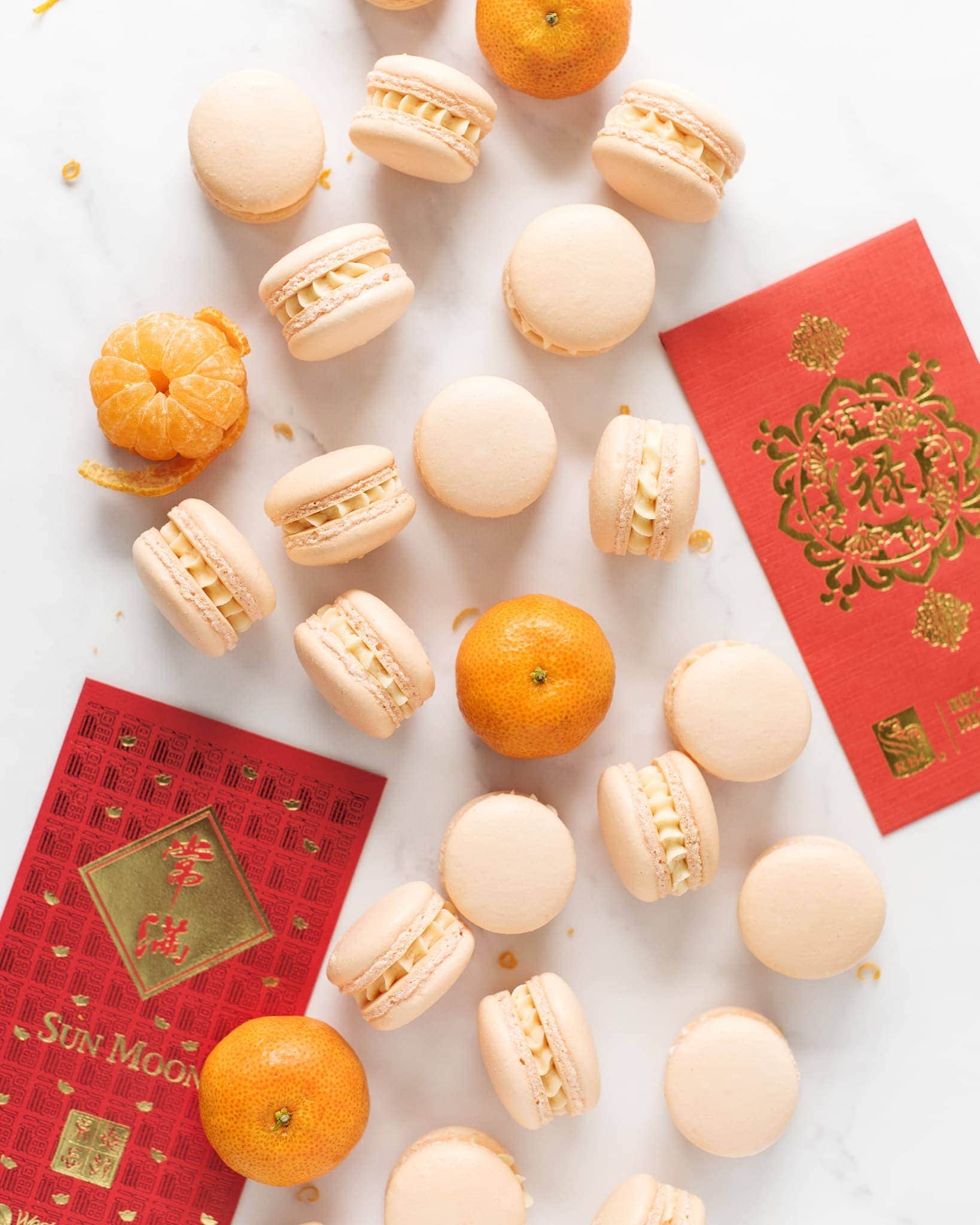 Mandarin orange macarons scattered on marble counter with Lunar New Year red envelopes