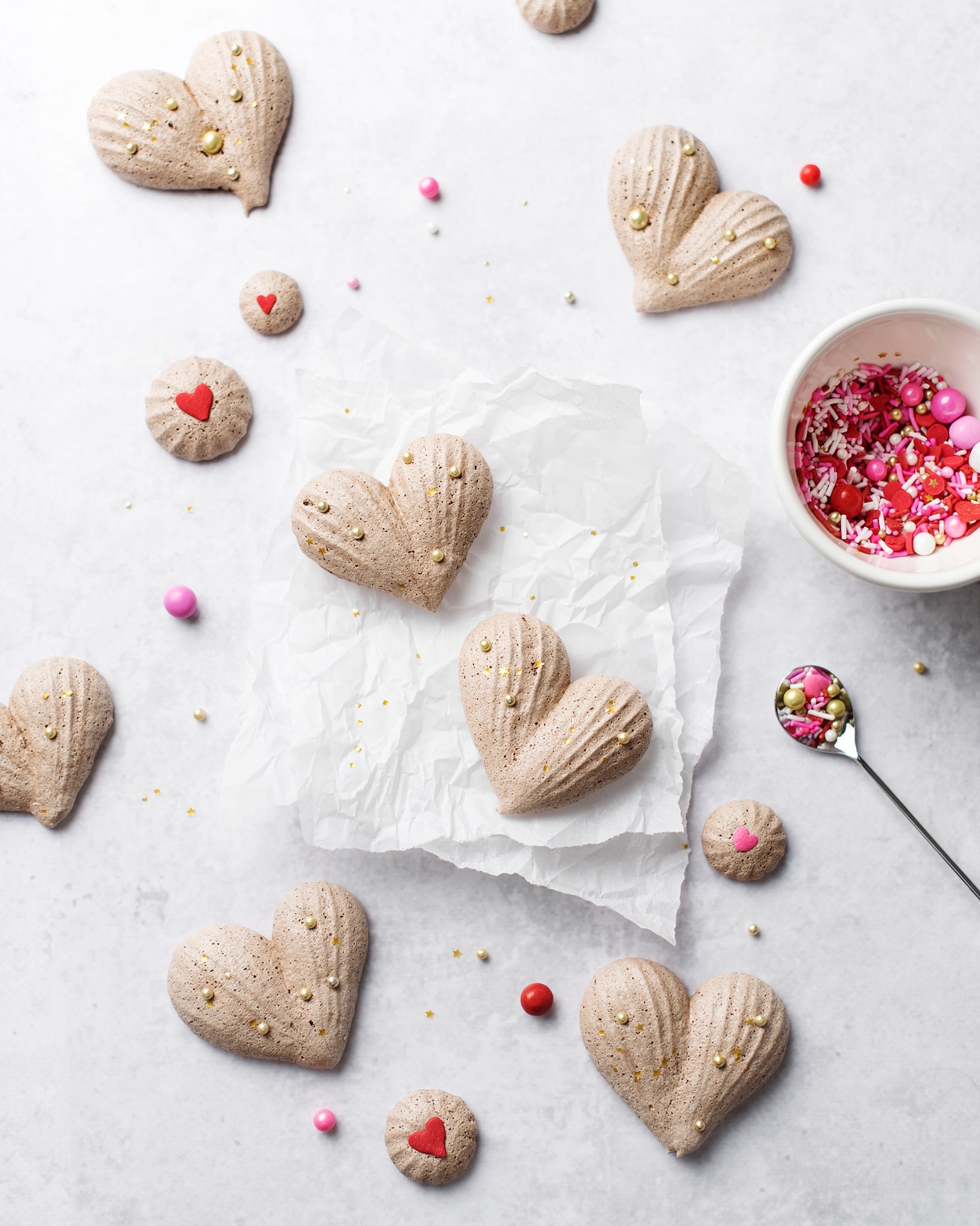 Heart-shaped chocolate meringues on parchment paper with a bowl of pink and red sprinkles
