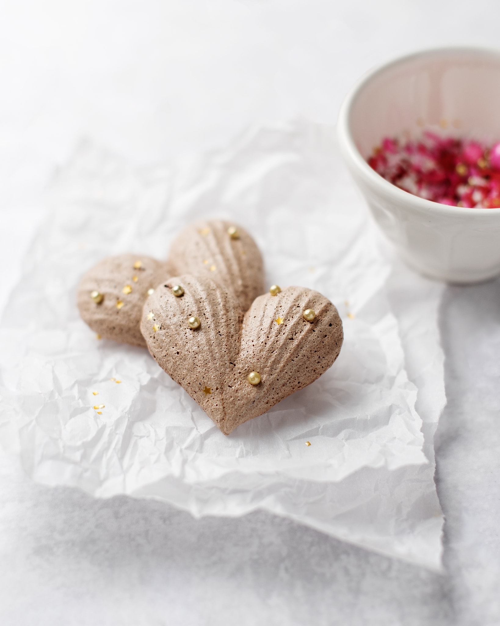 Two heart-shaped chocolate meringues on parchment paper with a bowl of sprinkles
