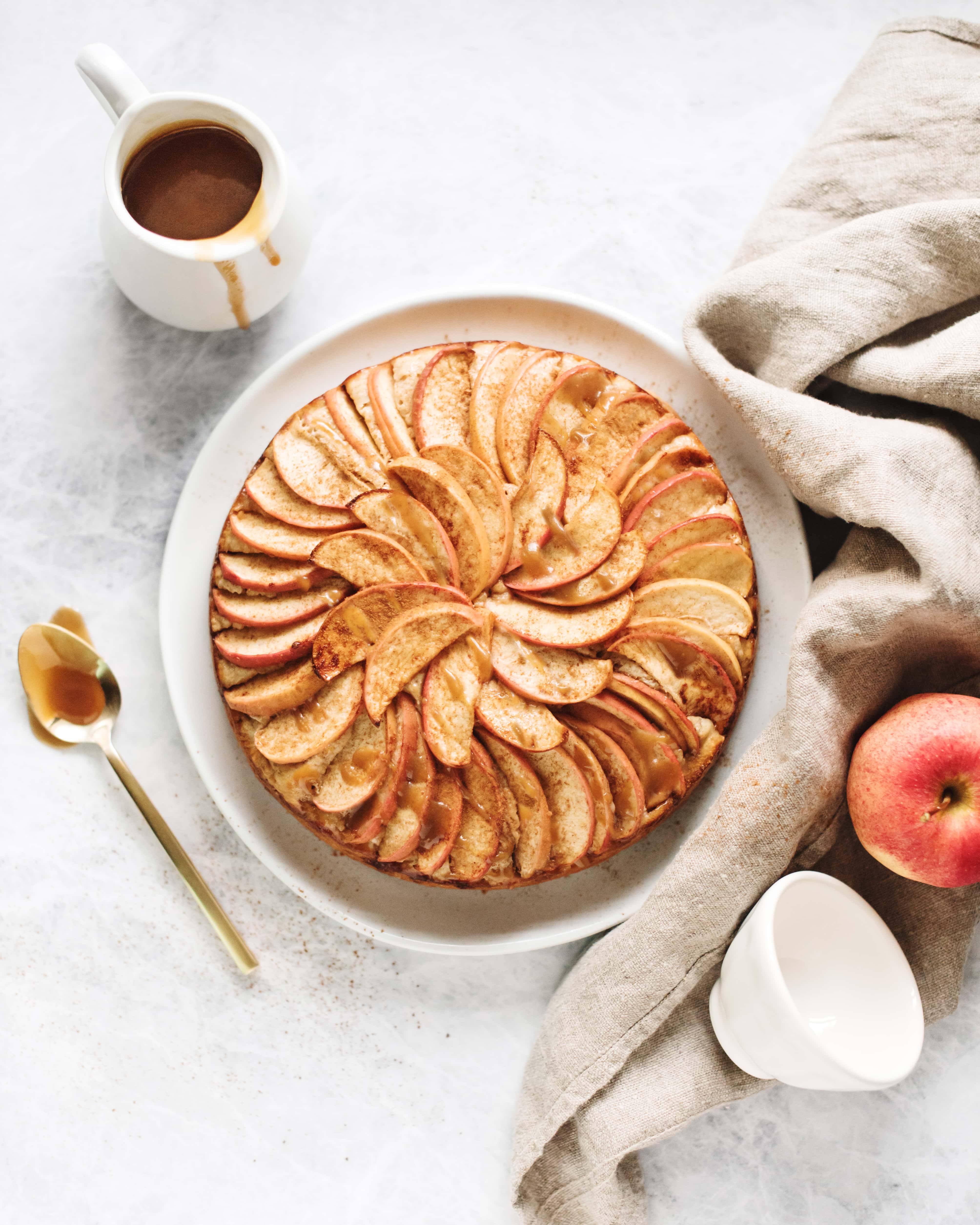Overhead shot of a spiced apple cake with salted maple caramel drizzle next to a brown linen and spoonful of caramel