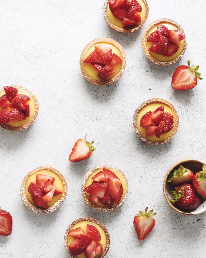 Overhead shot of lemon curd tartlets scattered on grey background with strawberries