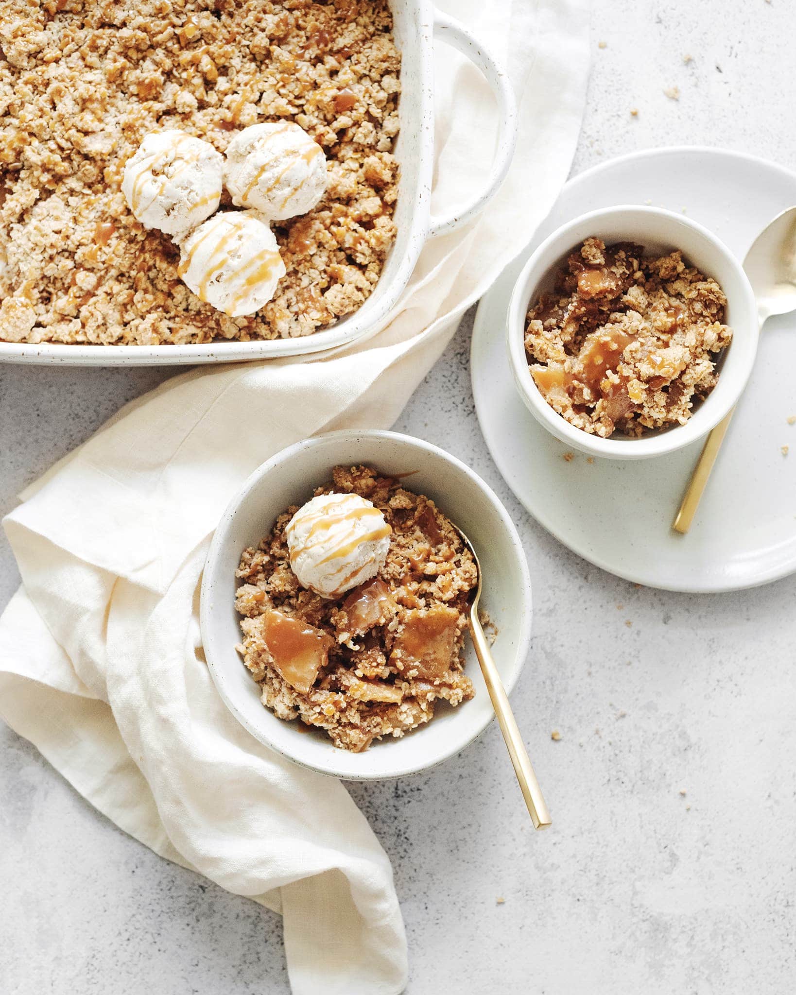 A baking dish and bowls of caramel apple crisp topped with ice cream