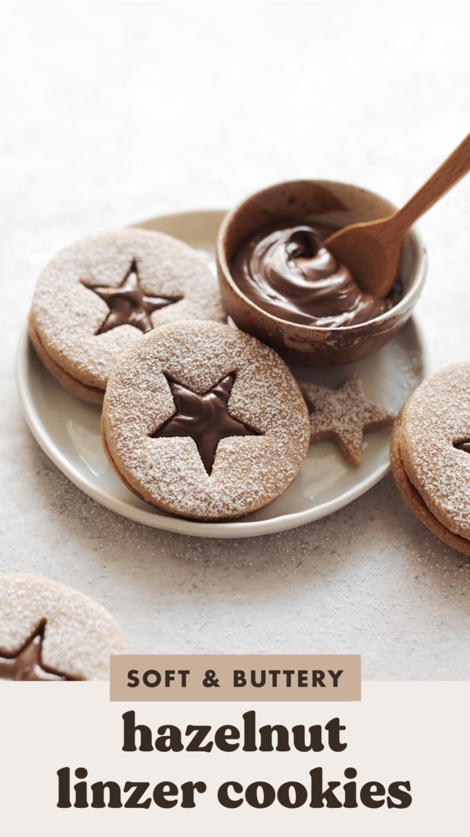 Two hazelnut linzer cookies on a plate with a small bowl of Nutella spread.