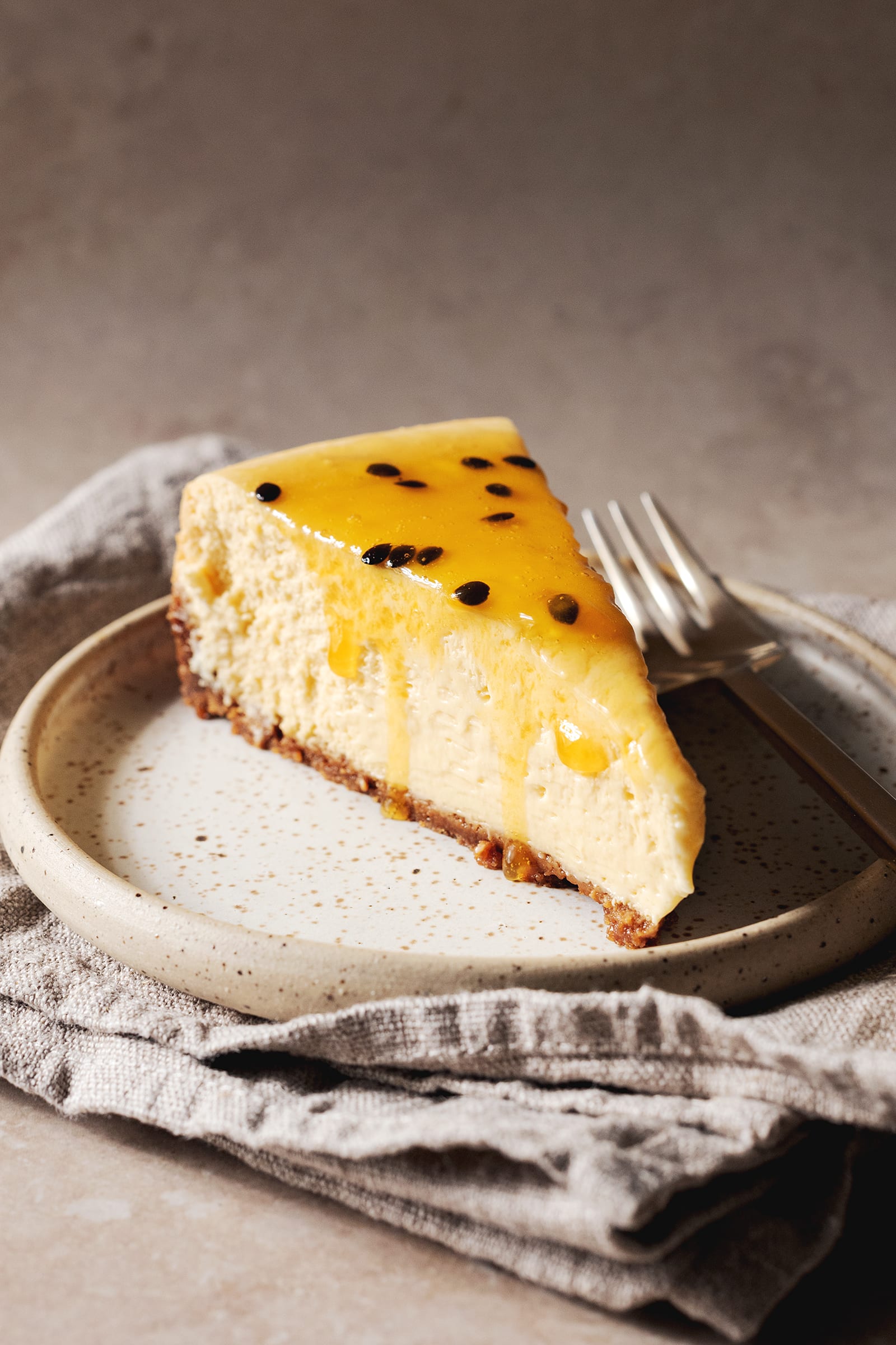 A slice of cheesecake with passionfruit puree dripping down the side on a speckled plate with a fork.