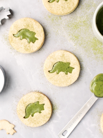 Matcha linzer cookies with triceratops cutouts on a grey background and sprinkled with matcha powder