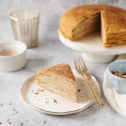 A slice of pumpkin crepe cake on a plate with the rest of the cake in the background