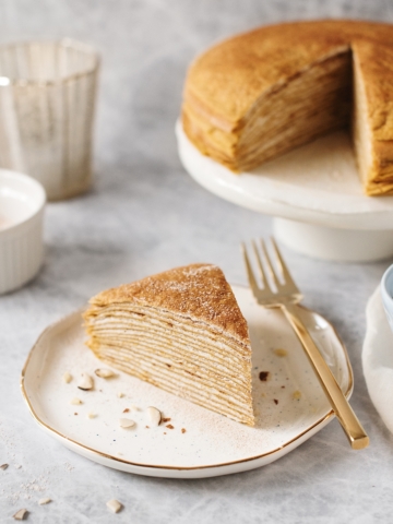 A slice of pumpkin crepe cake on a plate with the rest of the cake in the background