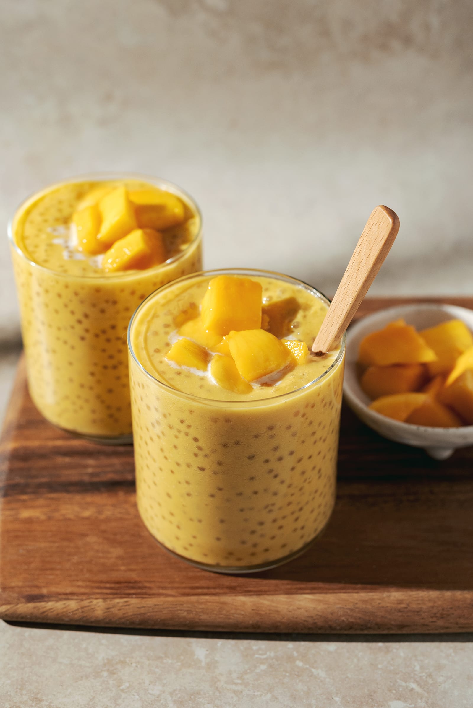 Two glasses of mango sago topped with mango chunks on a wooden platter.