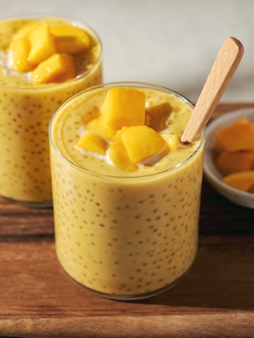 Two glasses of mango sago topped with mango chunks on a wooden platter.
