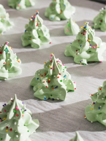 Meringue christmas trees covered in rainbow sprinkles on a baking sheet.