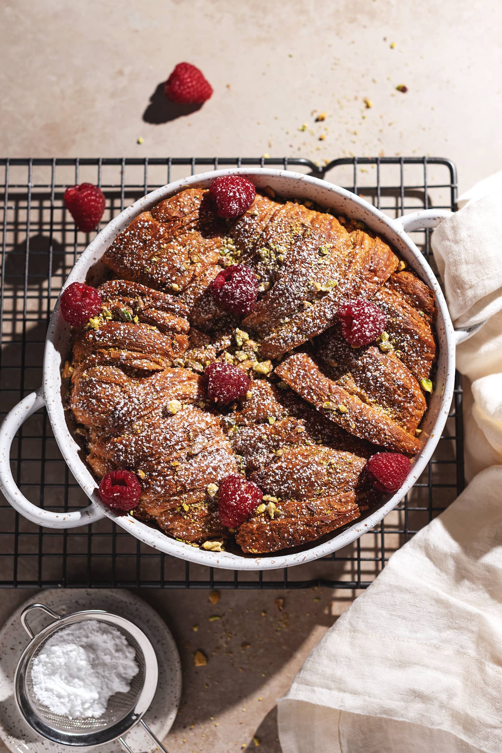 Baking dish filled with croissants sprinkled with raspberries, pistachios, and powdered sugar.