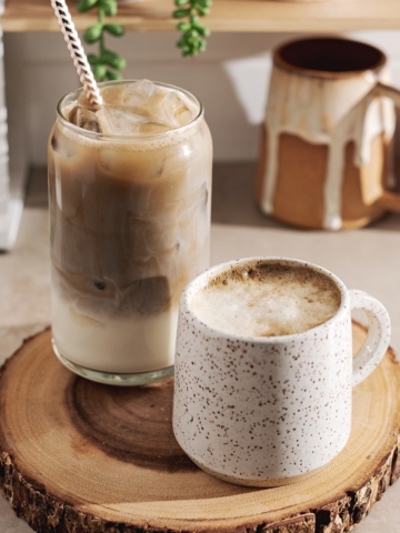 A glass of iced hojicha latte and a mug of hot hojicha latte sitting on wood platter.