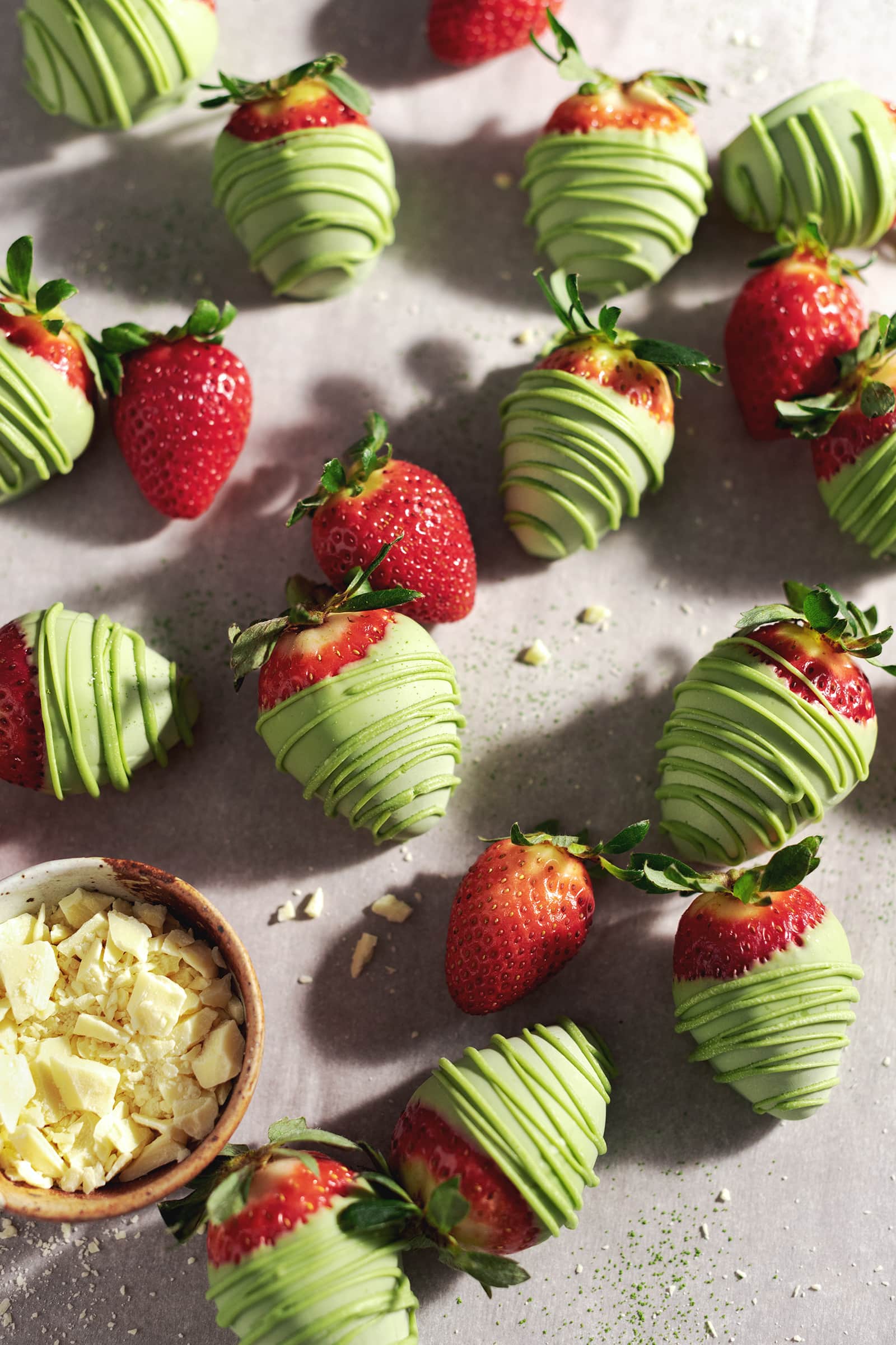 Matcha-covered strawberries scattered on table with fresh strawberries and a bowl of white chocolate.