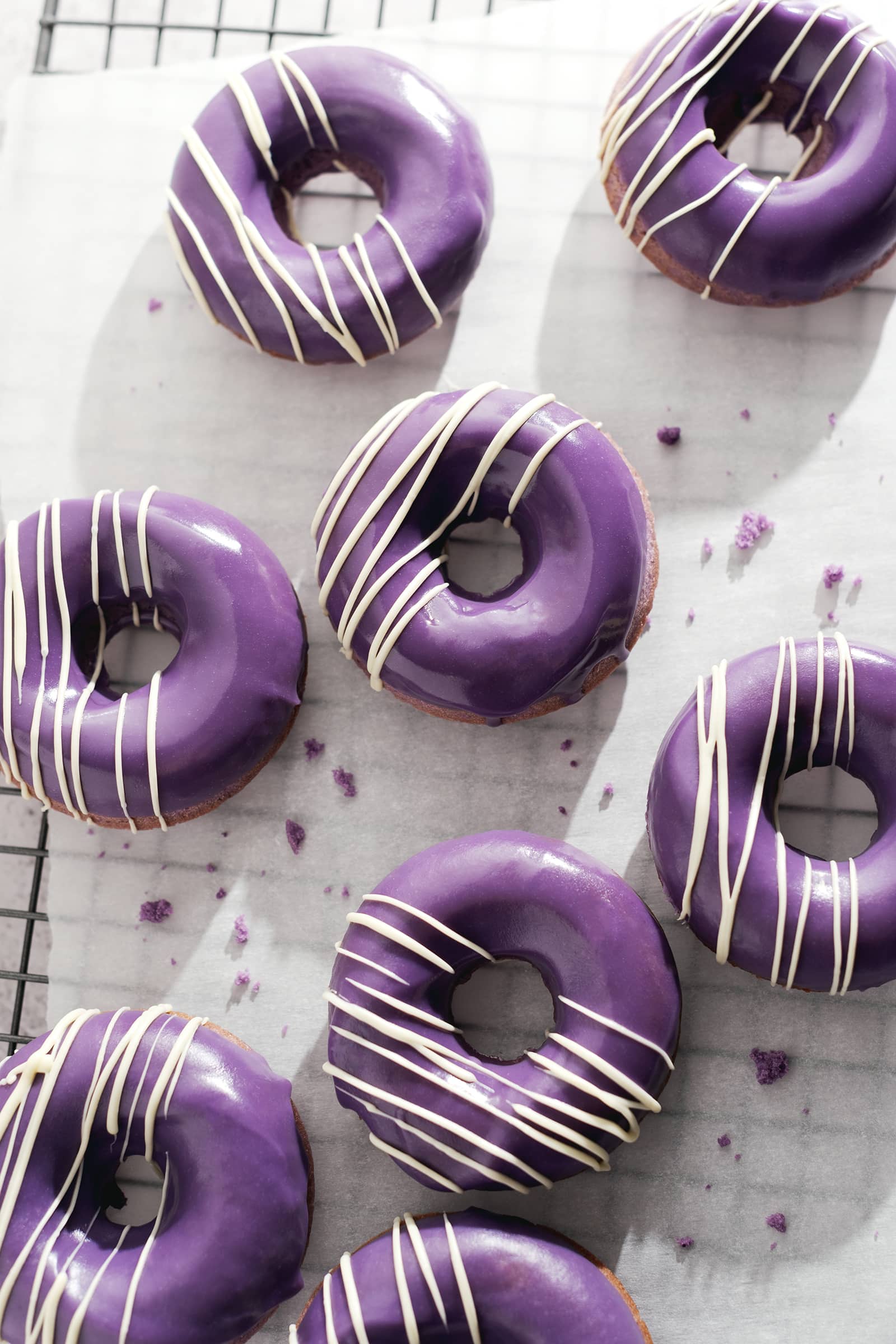 Ube donuts with white chocolate drizzles scattered on parchment paper on a wire rack.