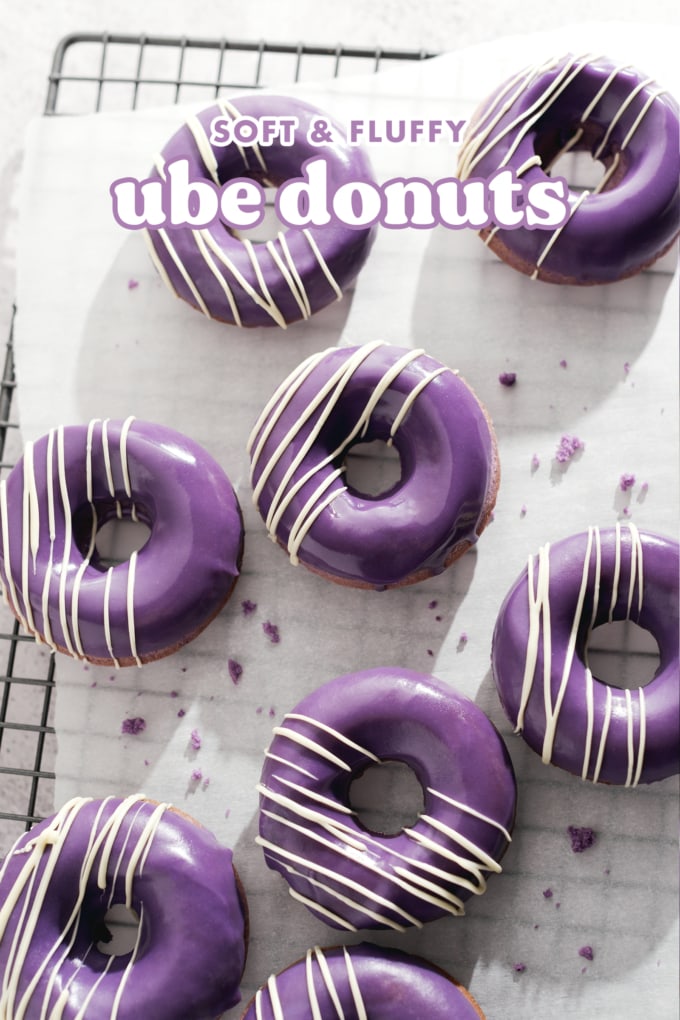 Ube donuts with white chocolate drizzles scattered on parchment paper on a wire rack.