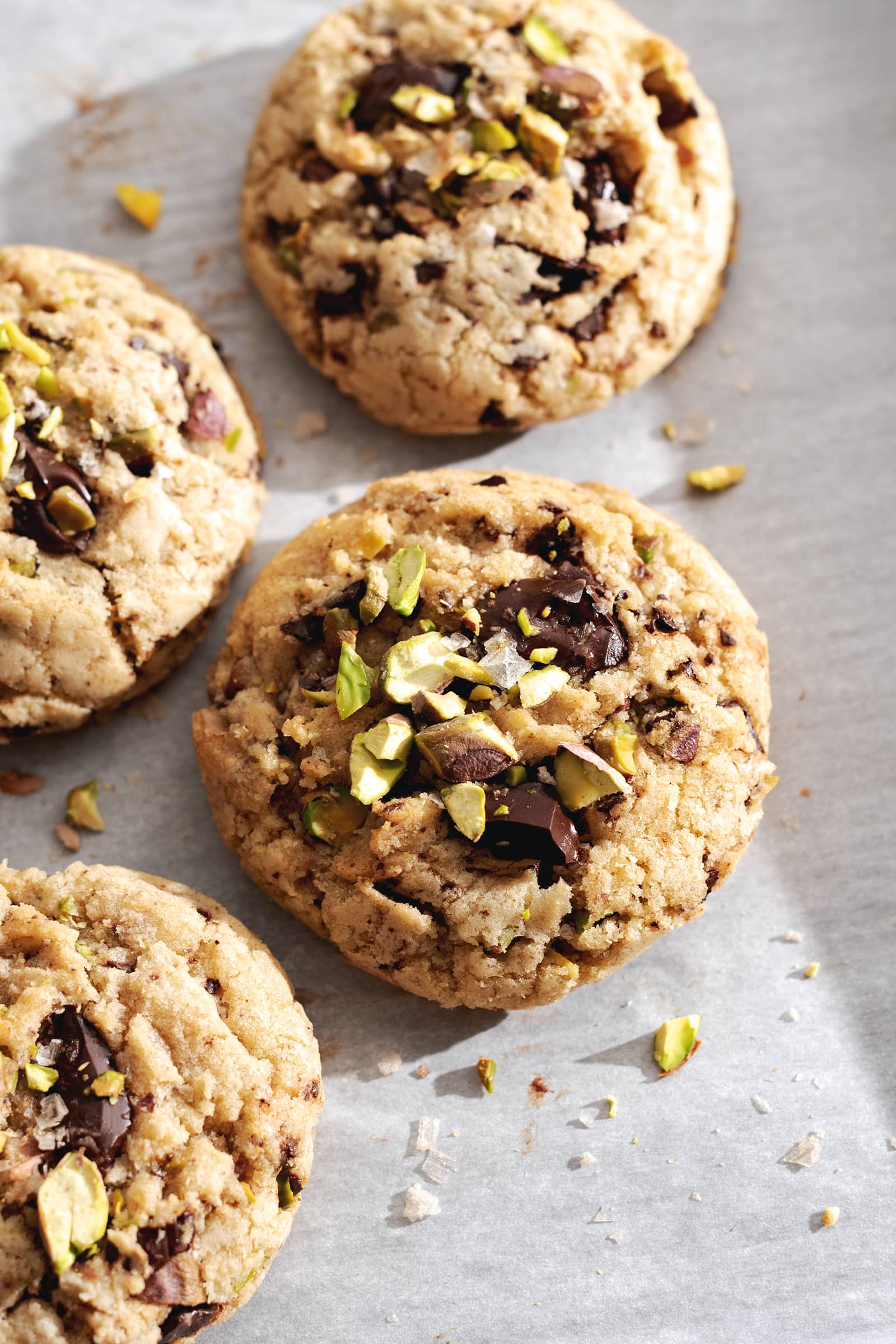 Four chocolate chip cookies with pistachios on top on parchment paper.