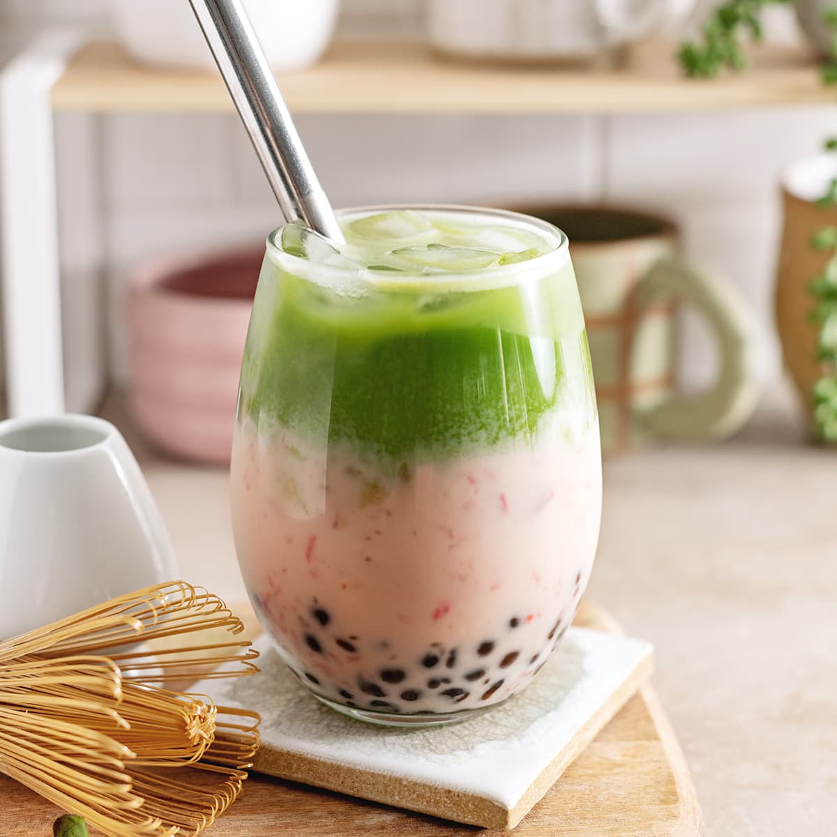 Strawberry matcha boba in a glass with straw and matcha whisk on the side.