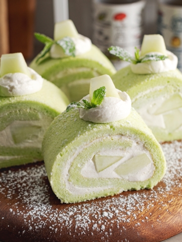 Four slices of honeydew roll cake sprinkled with powdered sugar on a wooden platter.