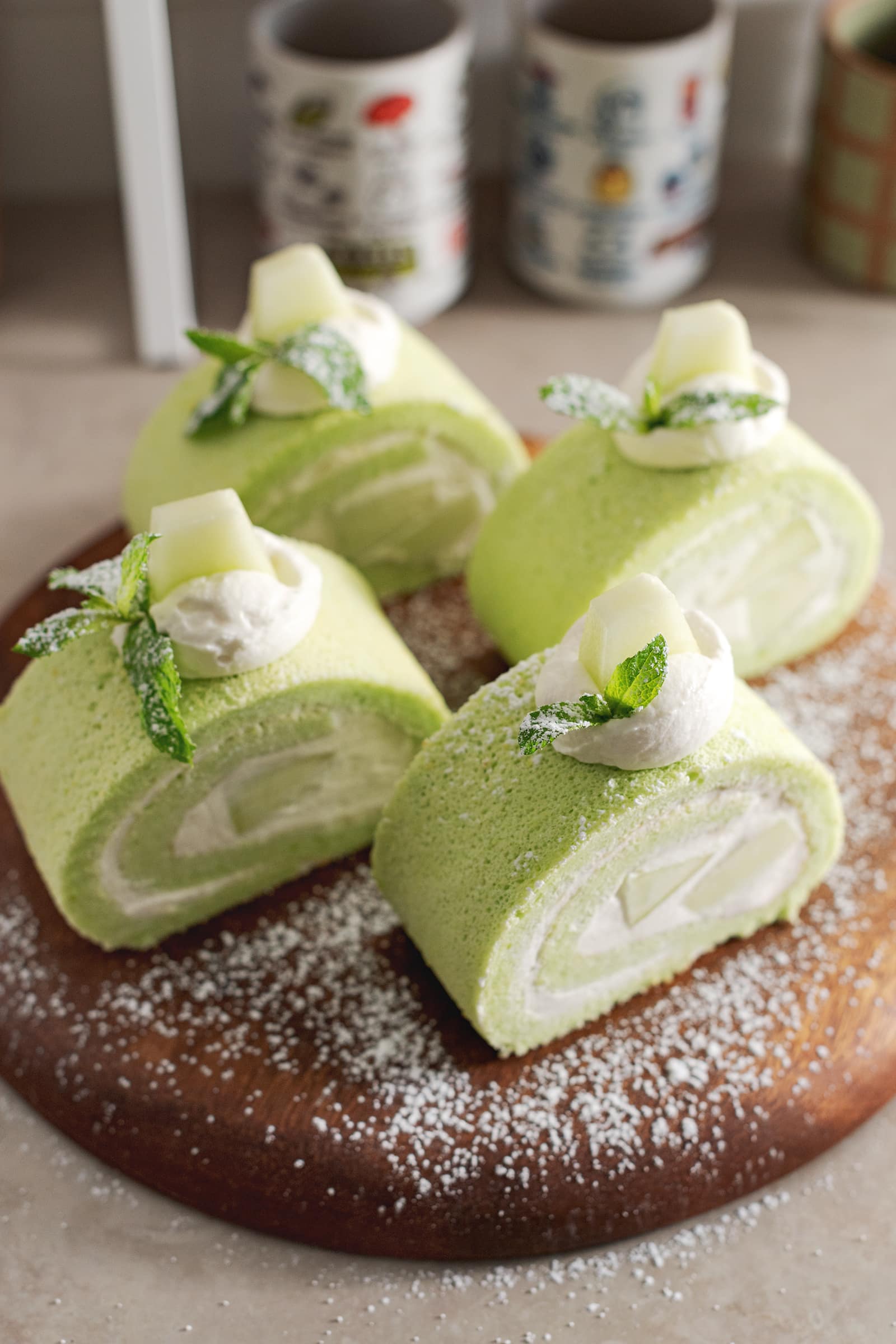 Four slices of honeydew roll cake sprinkled with powdered sugar on a wooden platter.