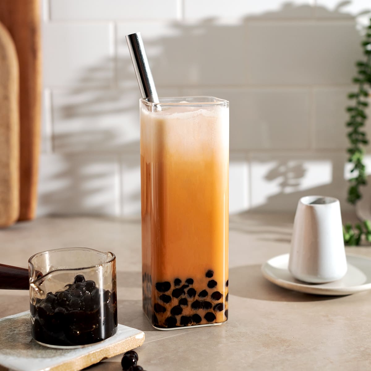 Thai milk tea boba in a rectangular glass with metal straw and small serving containers on either side.