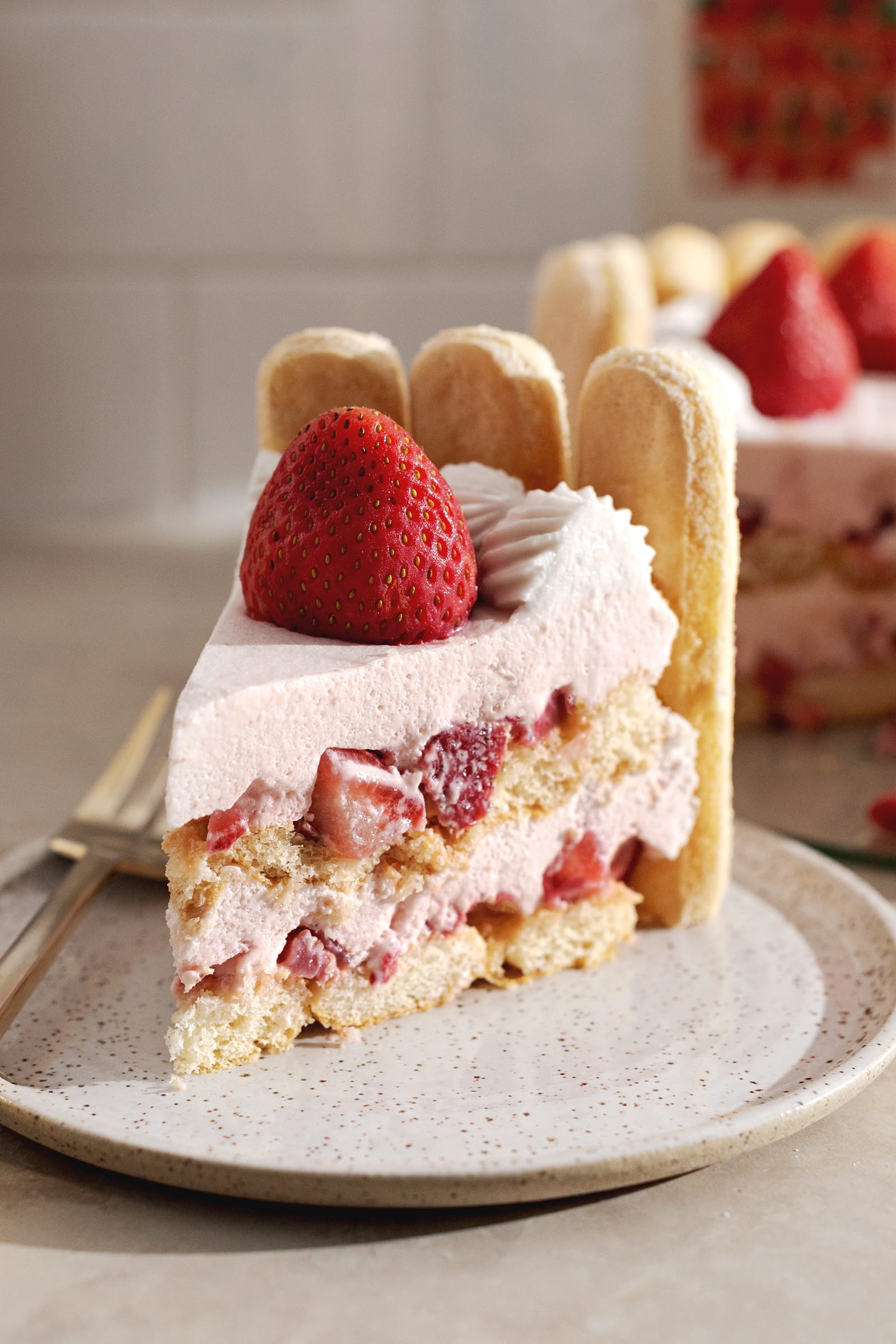 A slice of strawberry charlotte cake on a plate showing the layers and whole strawberry on top.