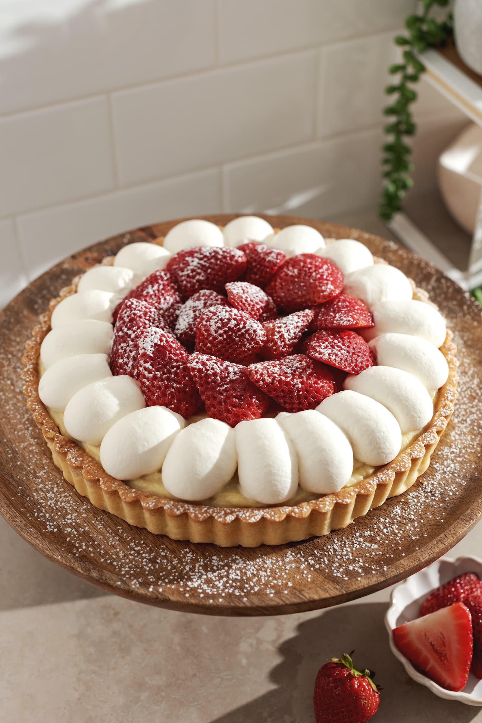Strawberry custard tart topped with dollops of whipped cream and layers of strawberries on a wooden plate.