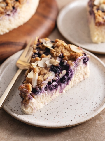 A slice of blueberry crumble cheesecake on a plate with a fork showing the layer of blueberries inside.