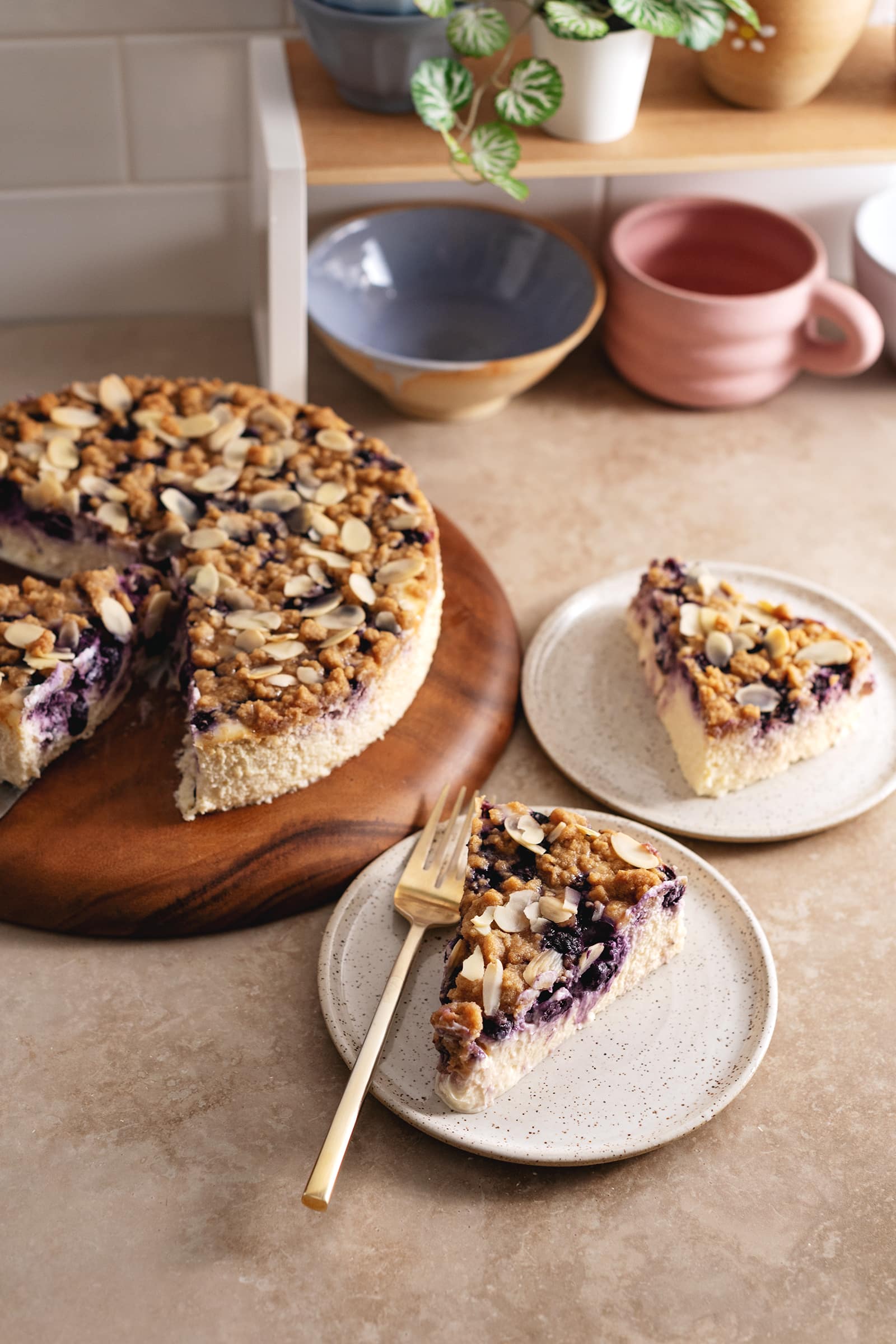 Slices of cheesecake with crumble topping on plates and the whole cheesecake on a wooden platter.