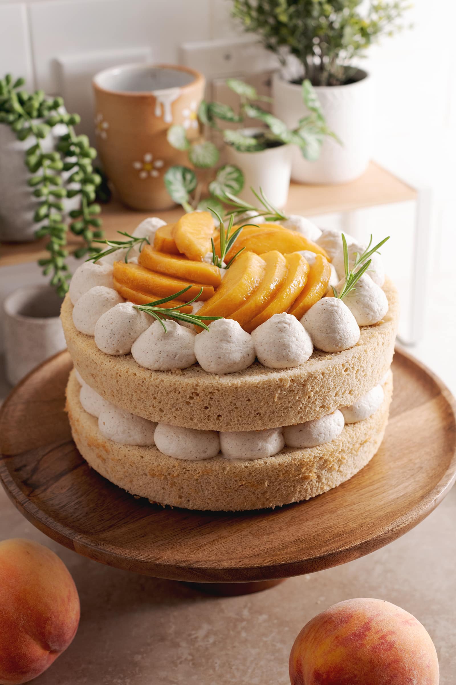 Two-layer peaches and cream cake on a wooden cake stand surrounded by peaches and plants.