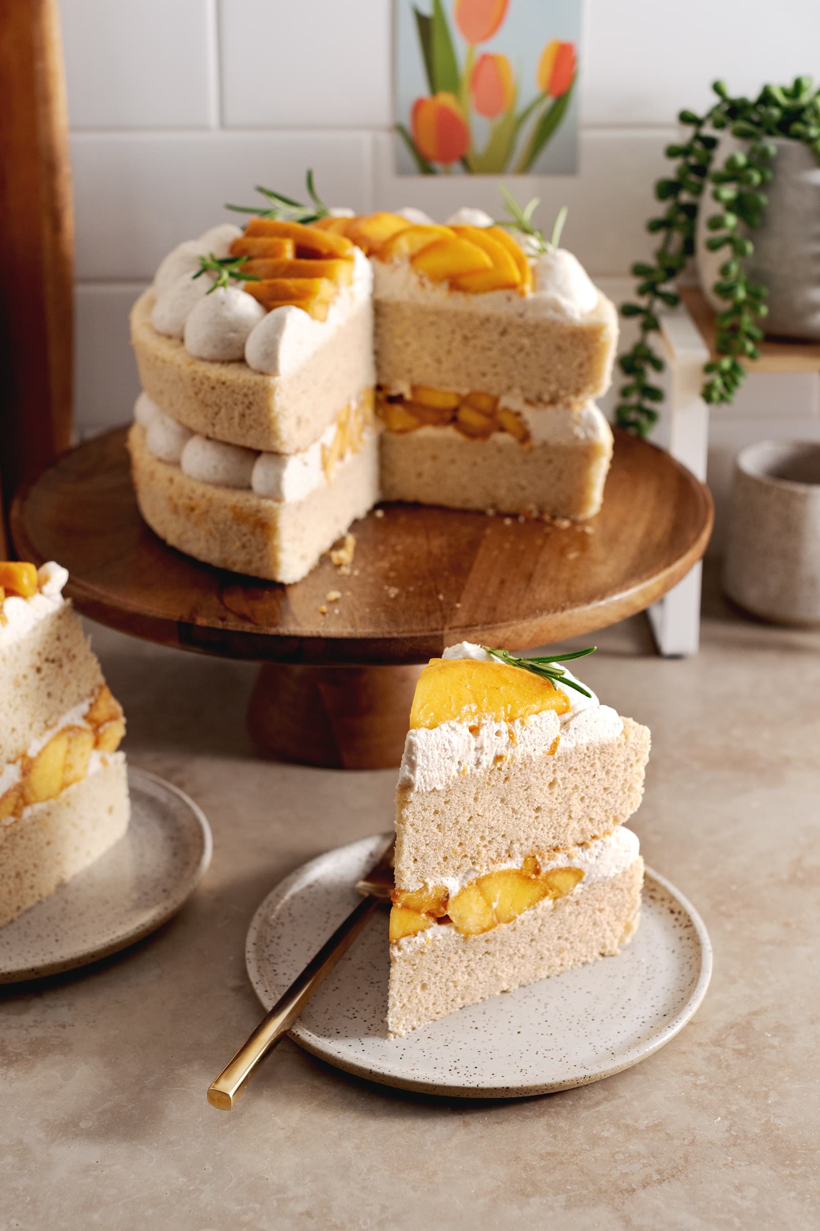 Two slices of peaches and cream cake on plates with the rest of the cake on a wooden cake stand behind them.