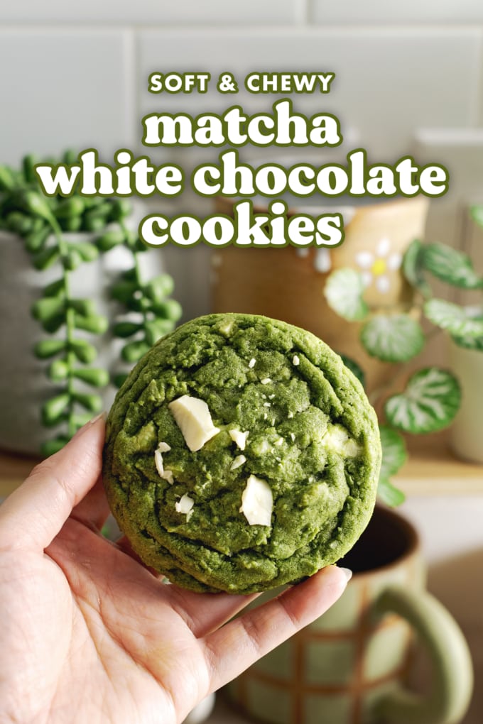 Hand holding a matcha cookie with white chocolate chunks in front of a shelf of mugs.