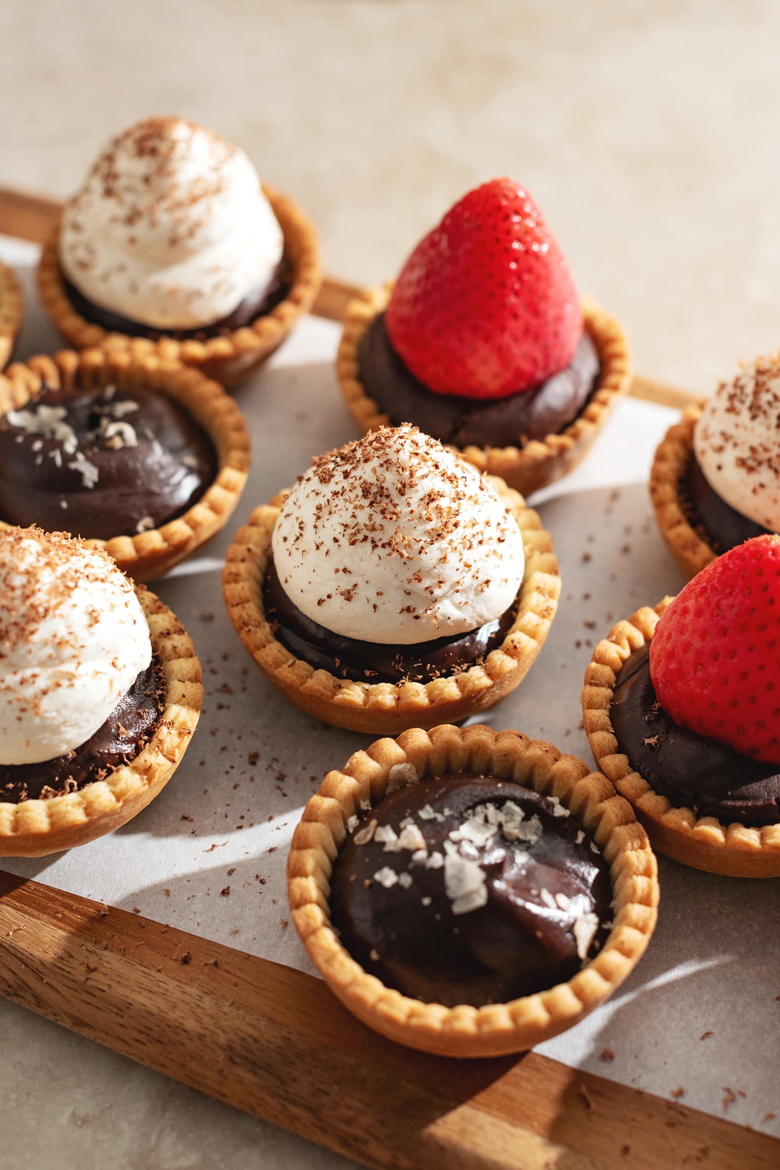 Mini chocolate tarts topped with salt flakes, whipped cream, and strawberries lined up on a wooden platter.
