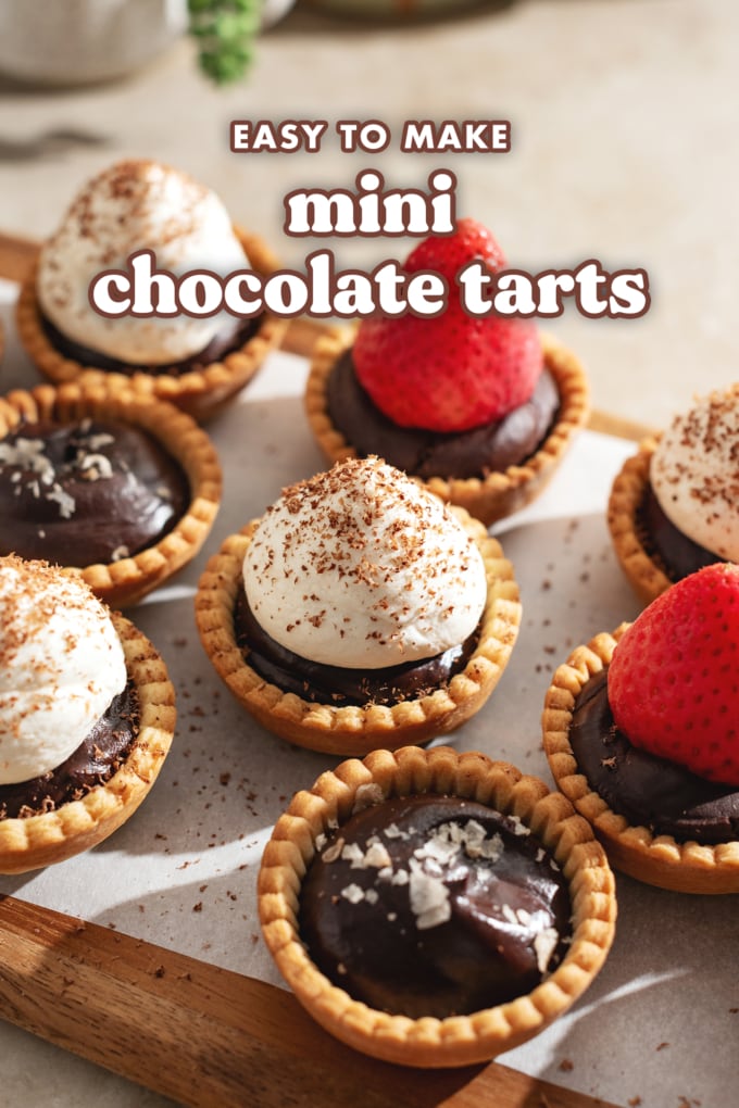 Mini chocolate tarts topped with salt flakes, whipped cream, and strawberries lined up on a wooden platter.