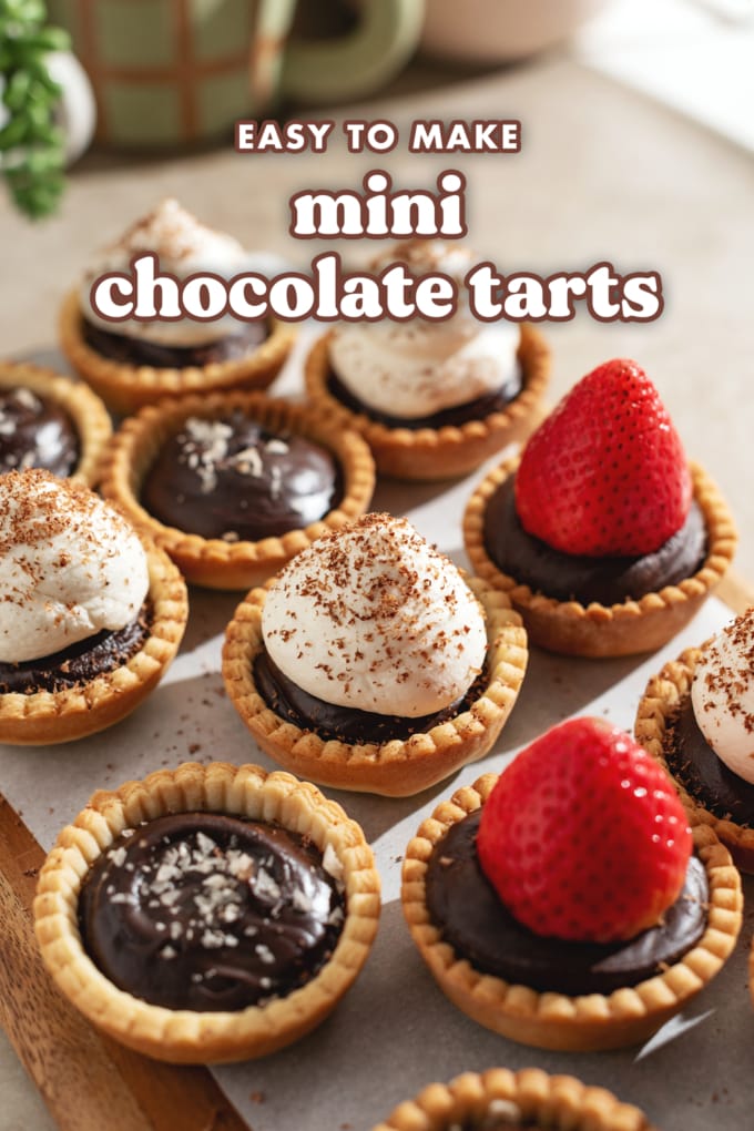 Mini chocolate tarts topped with salt flakes, whipped cream, and strawberries lined up on a wooden platter.