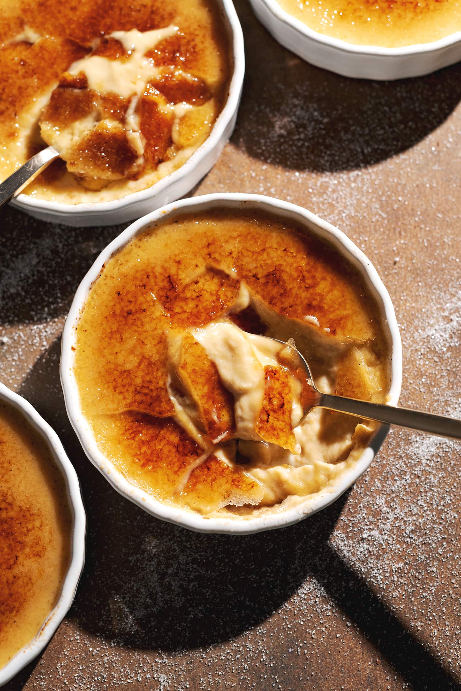 Ramekins of crème brûlée cracked with spoons to show the custard inside.