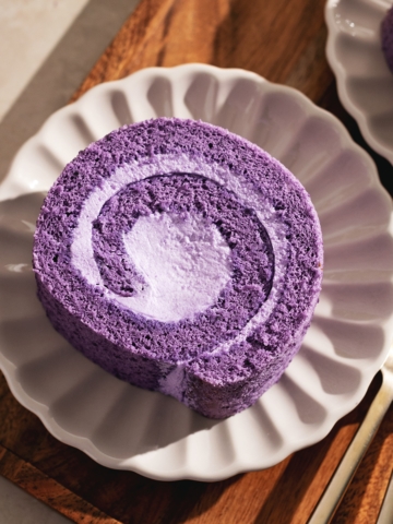 Top down view of a slice of ube roll cake on a scalloped plate on a wooden tray.