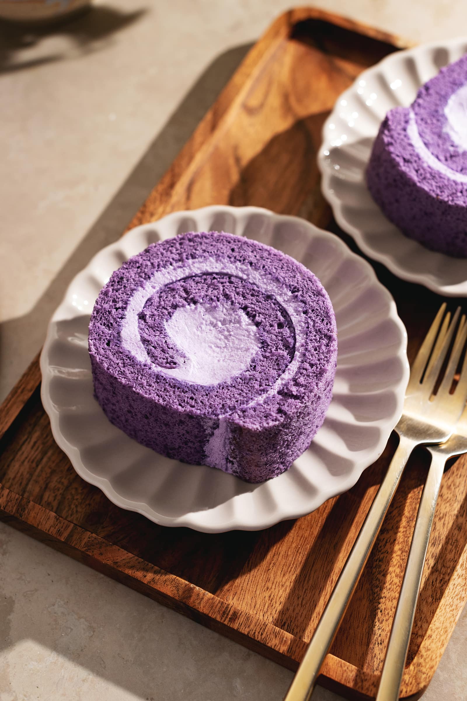 A slice of ube roll cake on a scalloped plate on a wooden tray.