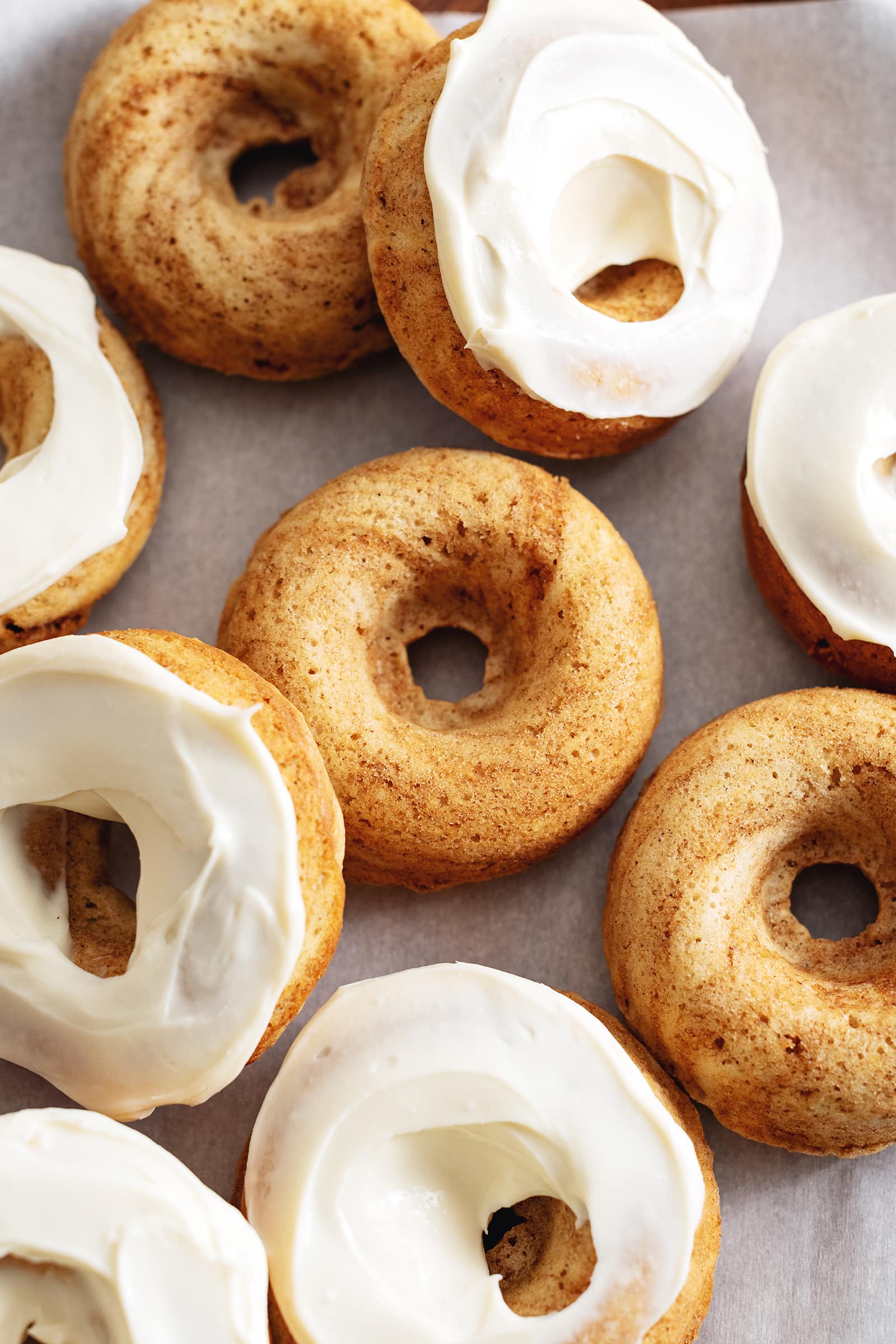 Cinnamon roll donuts scattered on parchment paper, some with cream cheese frosting, some without.