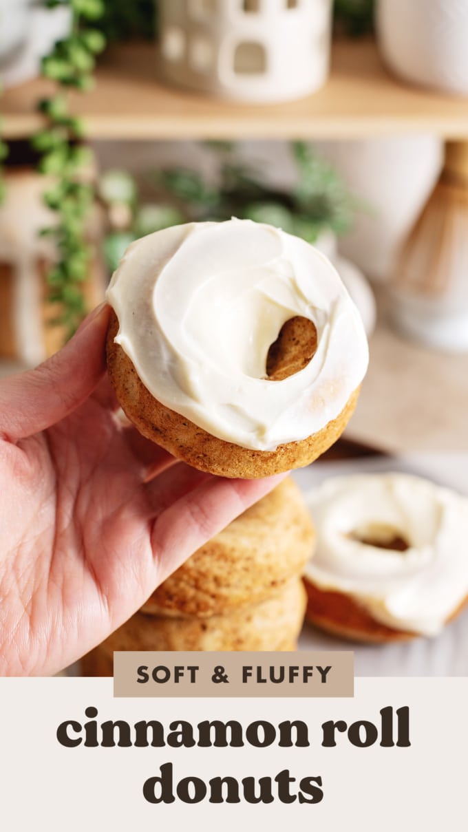 A hand holding a cinnamon roll donut frosted with cream cheese icing.