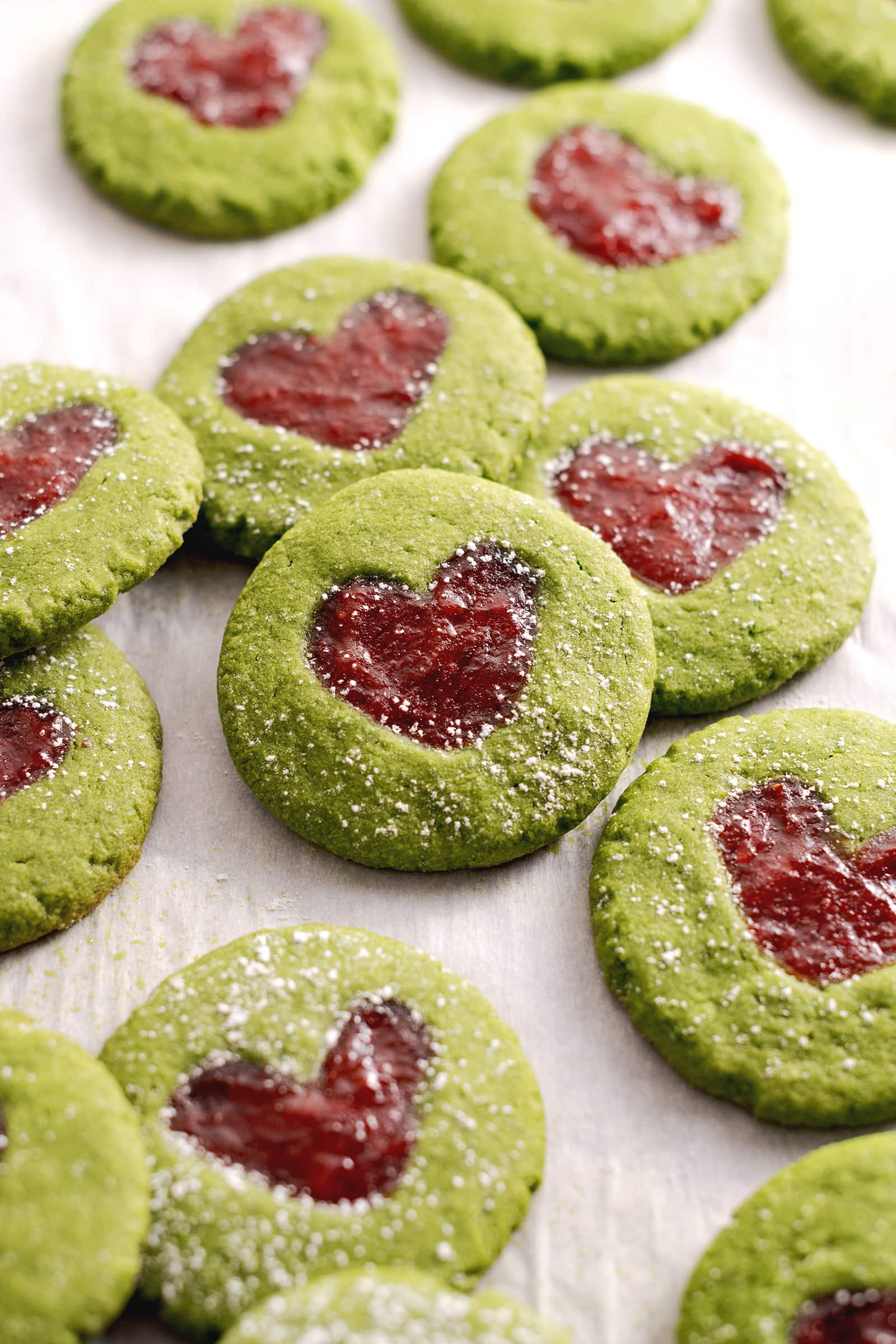 Matcha thumbprint cookies leaning against other cookies with strawberry jam in the shape of a heart.