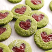 Matcha thumbprint cookies leaning against other cookies with strawberry jam in the shape of a heart.
