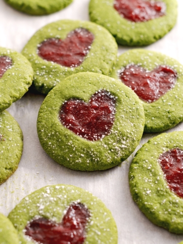Matcha thumbprint cookies leaning against other cookies with strawberry jam in the shape of a heart.