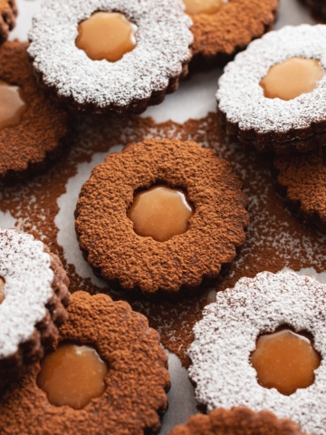 Several salted caramel chocolate linzer cookies, some dusted with cocoa powder and some with powdered sugar, scattered on parchment paper.