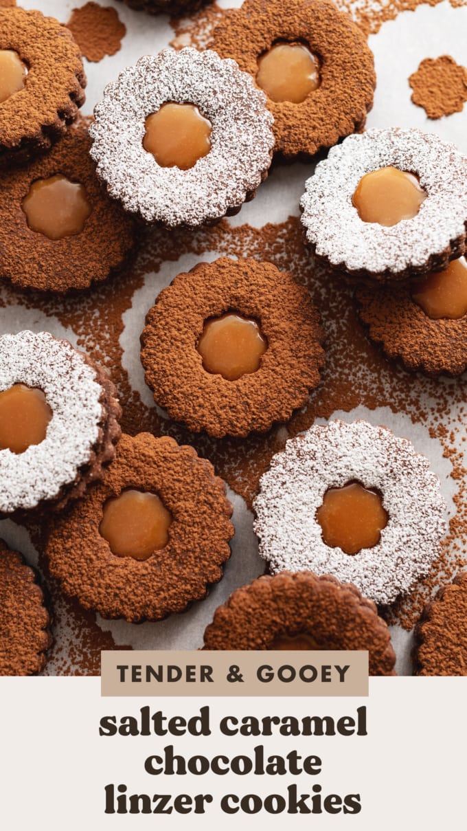 Several salted caramel chocolate linzer cookies, some dusted with cocoa powder and some with powdered sugar, scattered on parchment paper.